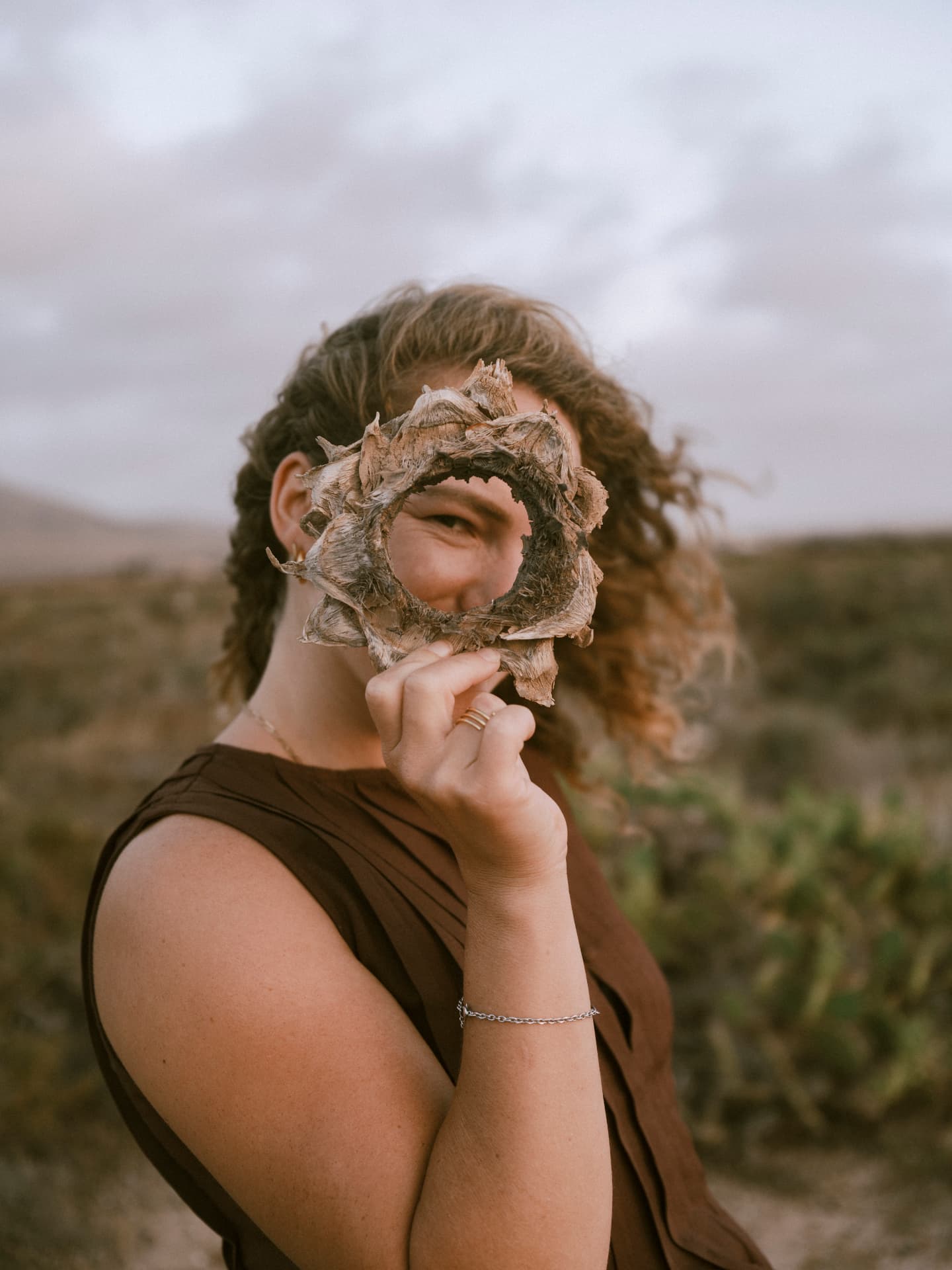 Elena holding a dried seed pod in front of her eye in a coastal landscape