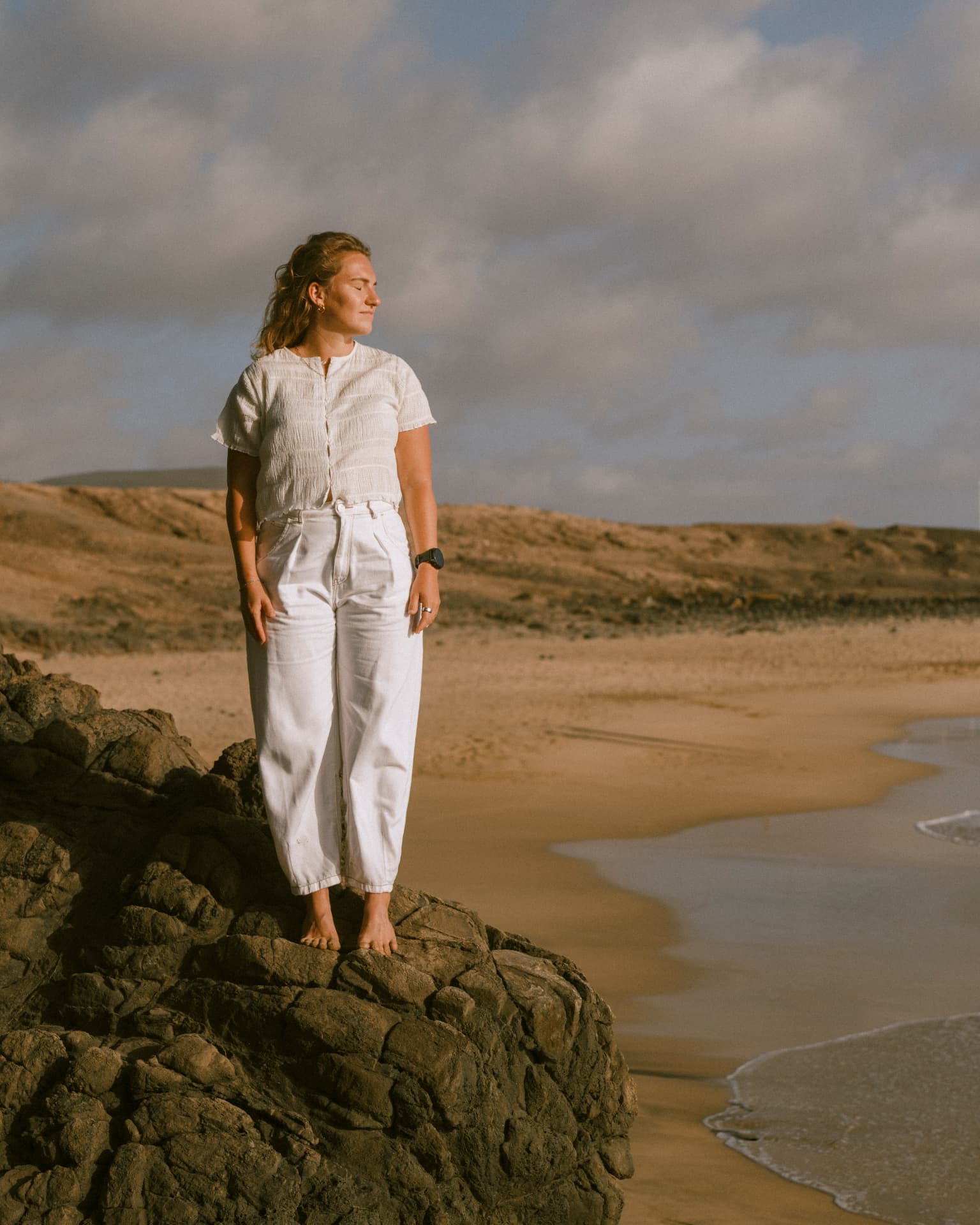 Elena standing barefoot on a rock above a curving sand beach